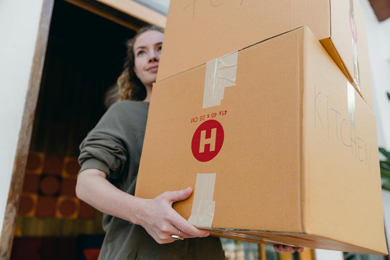 From below of young slim woman carrying stack of heavy carton boxes with kitchen stuff and looking away while moving out of old house