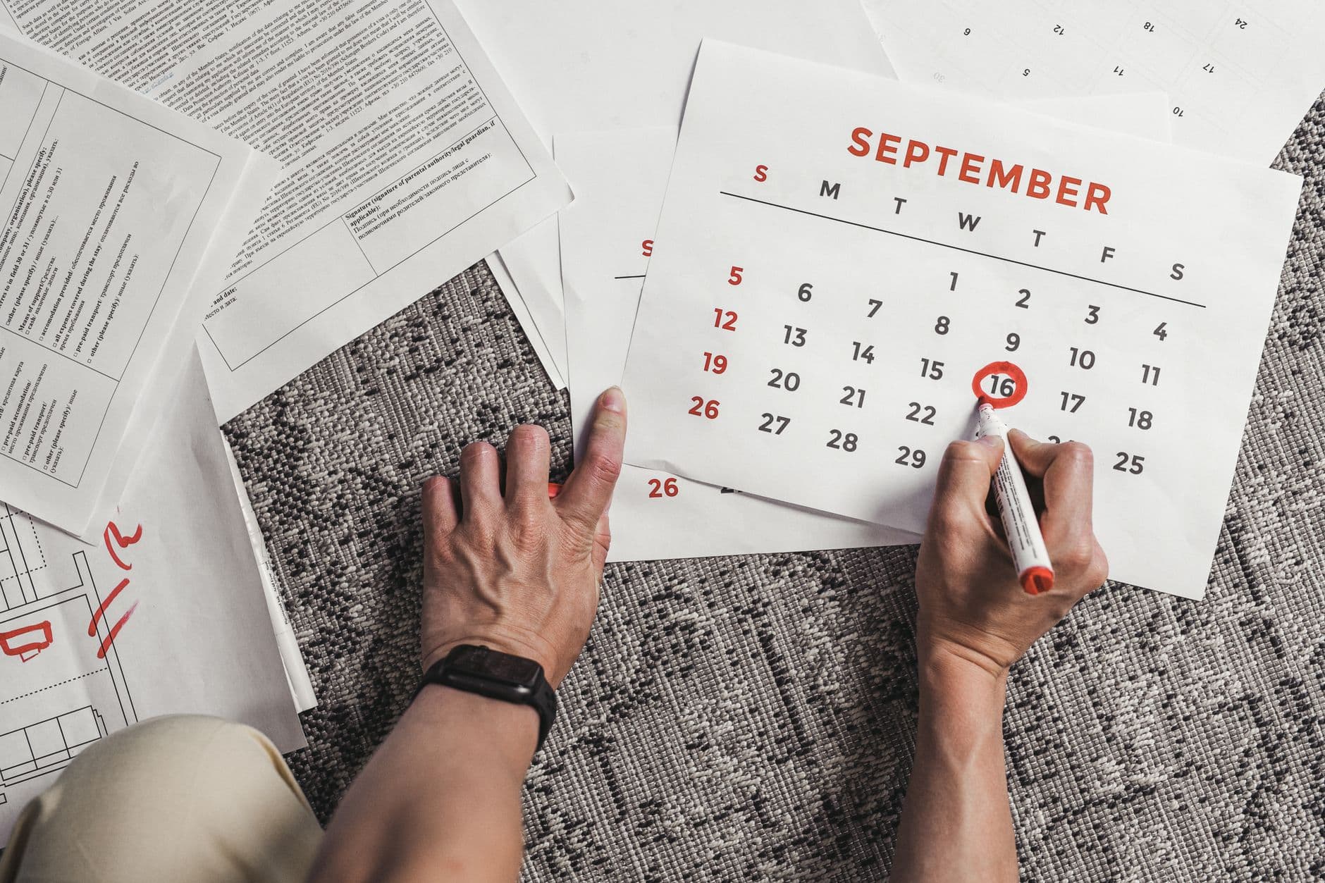 Hands holding a calendar with September dates encircled, surrounded by documents on a carpet.